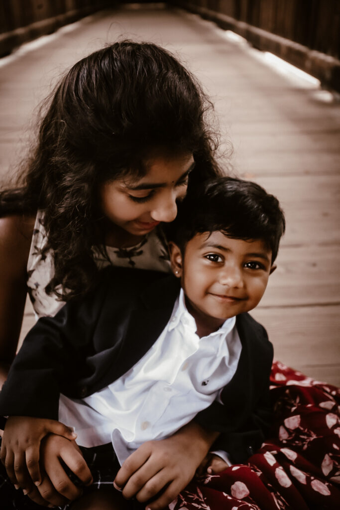 Sister holding her little brother in her lap while smiling at him as he smiles at the camera