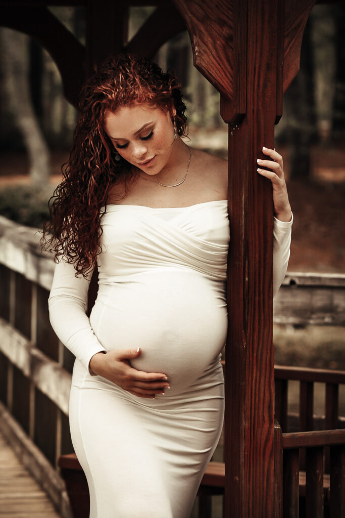 Pregnant mom holding her belly while in a long while dress and holding onto a wooden pole looking at he belly