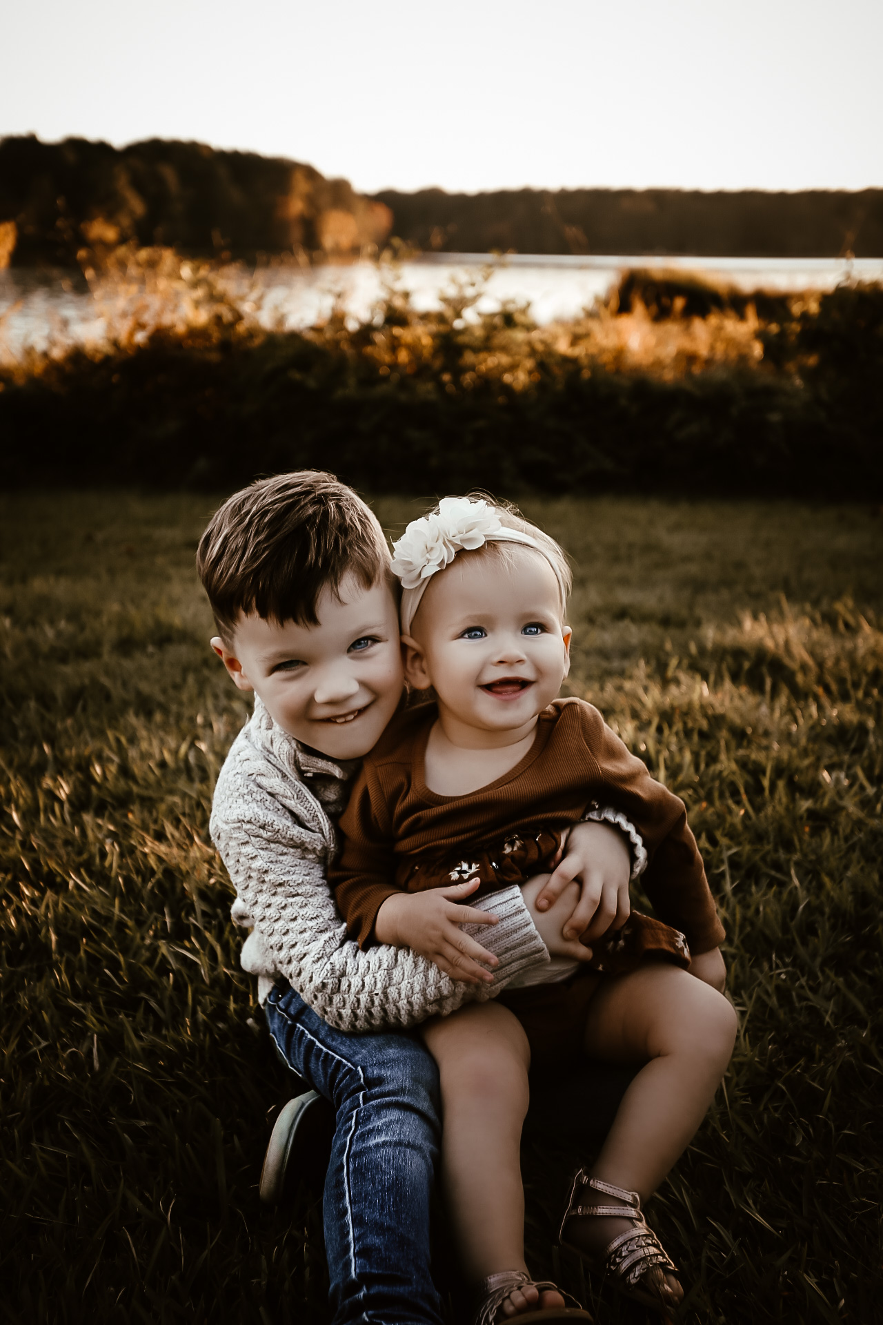 Toddler boy holding his baby sister while sitting on the ground and both smiling at the camera