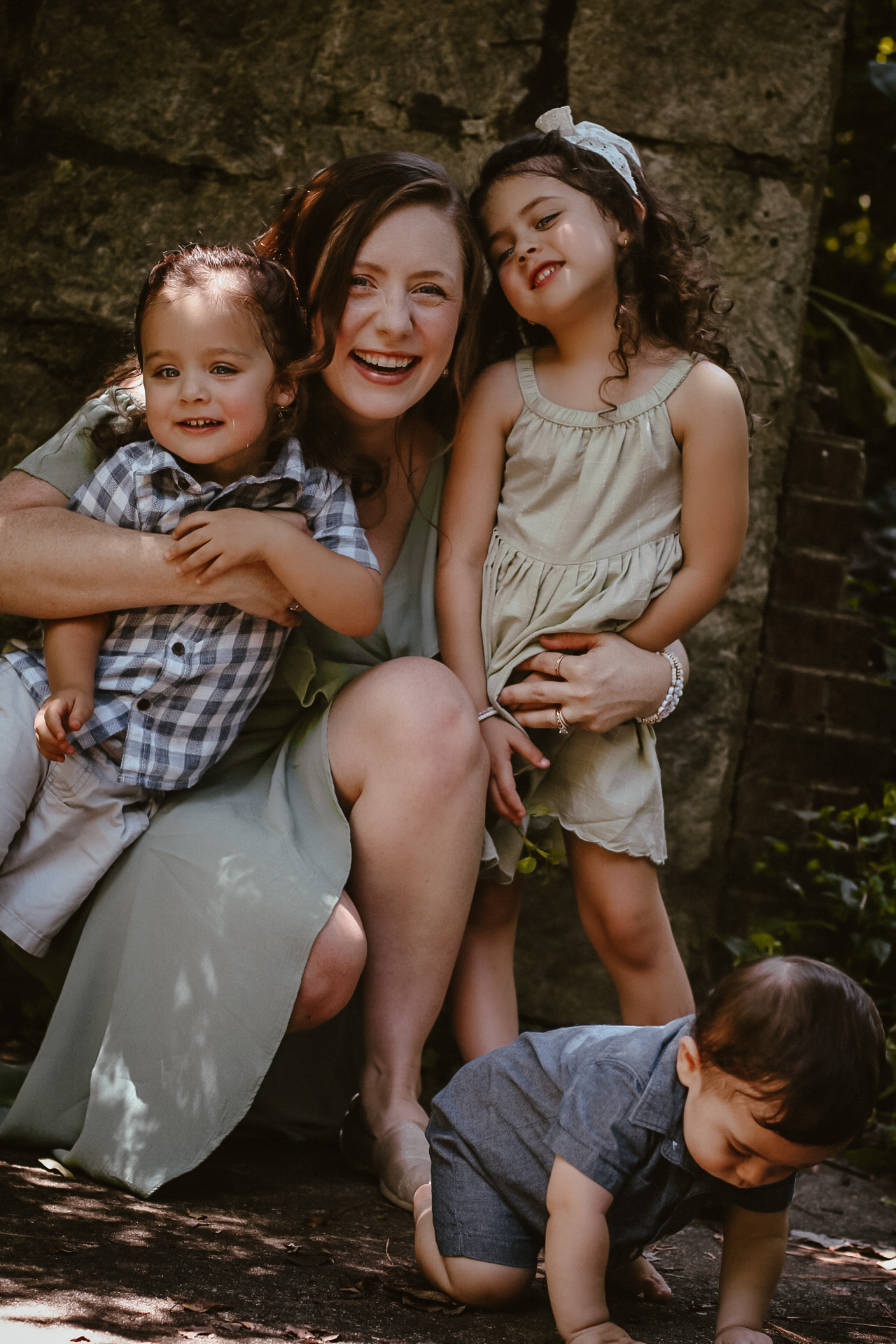 Mom holding her 3 babies while all smiling at the camera and baby on the ground crawling