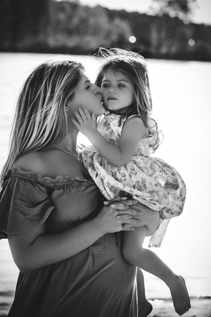 Mom holding her young daughter while kissing her on the cheek and daughter embracing her mom and looking off into the distance