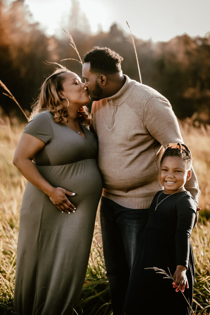 Family of 3 pregnant mom kissing her husband and young daughter smiling at the camera