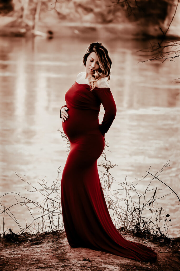 Pregnant mom standing by a lake in a red dress while holding her belly and looking down at the ground