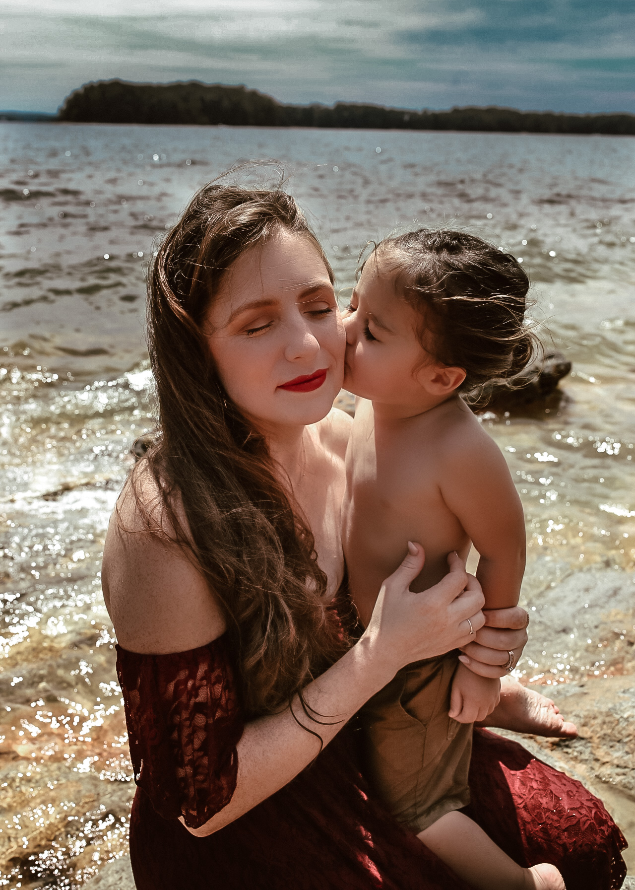 Mom and son sitting on a rock in the water at Lake Lanier while son is kissing mom and the cheek with their eye's closed