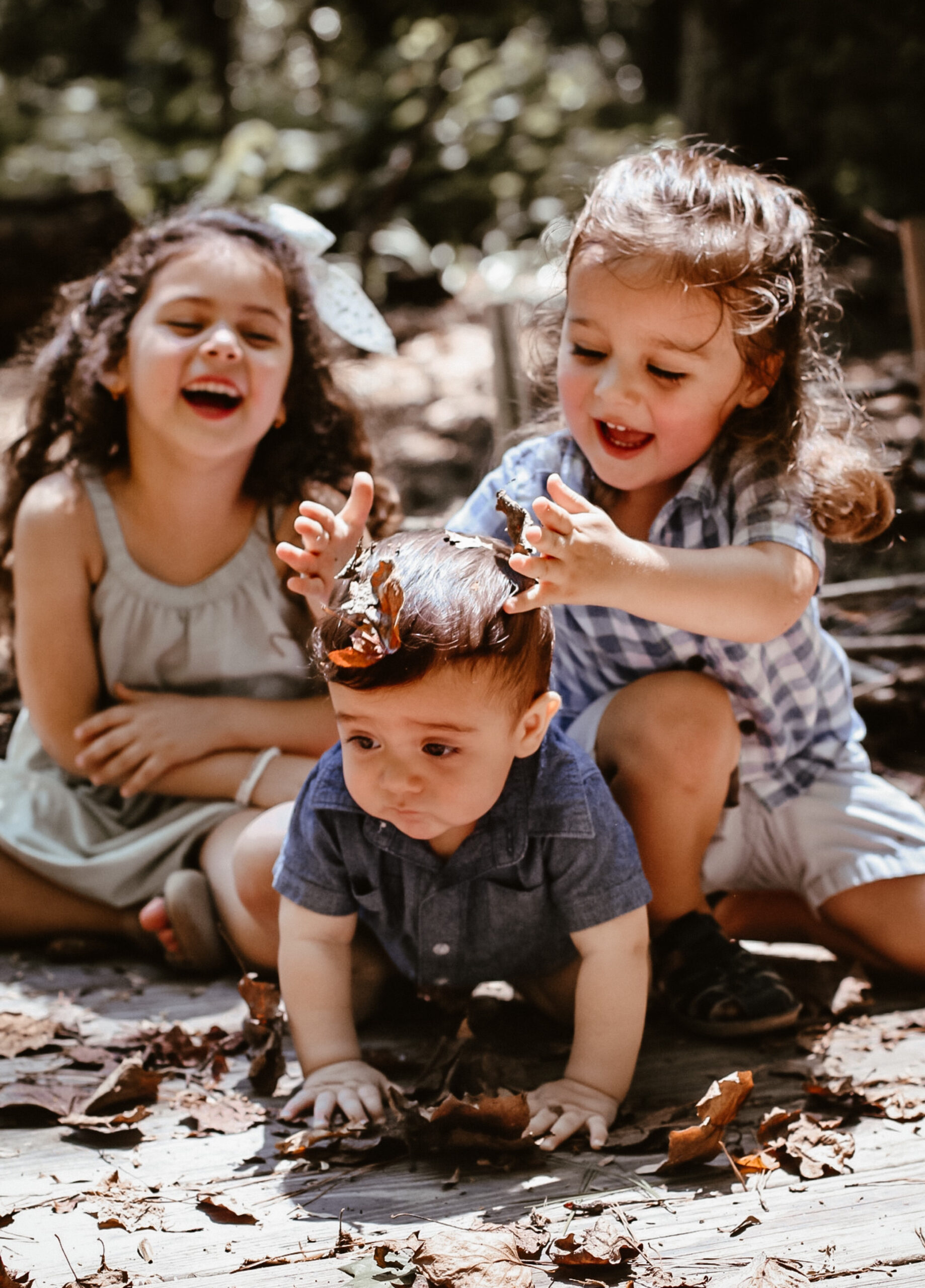 Three sibling laughing and playing together while the oldest son puts leaves on the youngest sons head and daughter laughing in the background