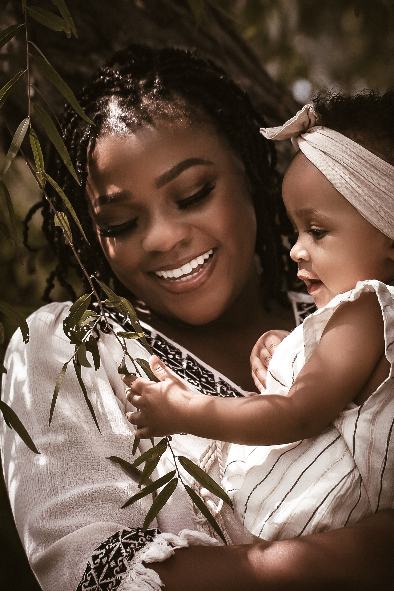 Mom holding her young baby while smiling and playing with a leaf on the tree