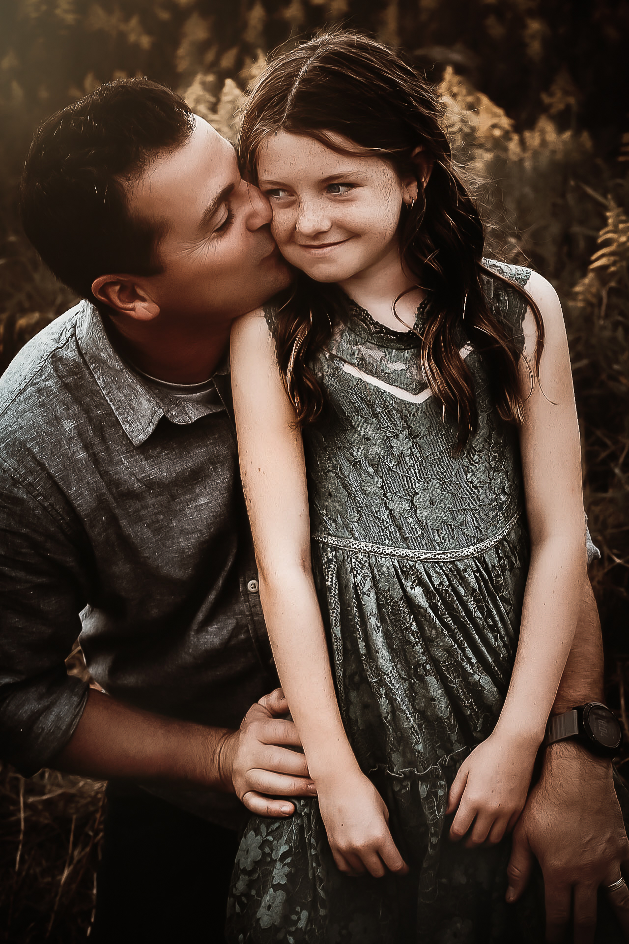 Dad kneeling on the ground while daughter is sitting on his lap and dad is kissing her on the cheek