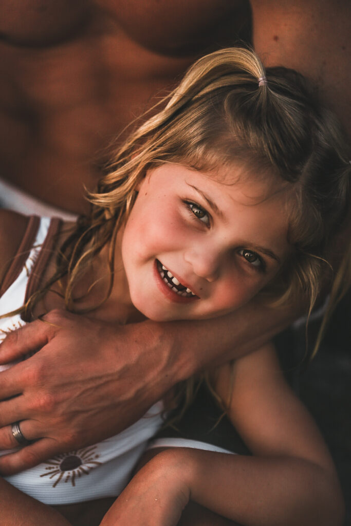 Little girl laying on her dads lap at the beach while smiling at the camera - located at St. Andrew's State Park