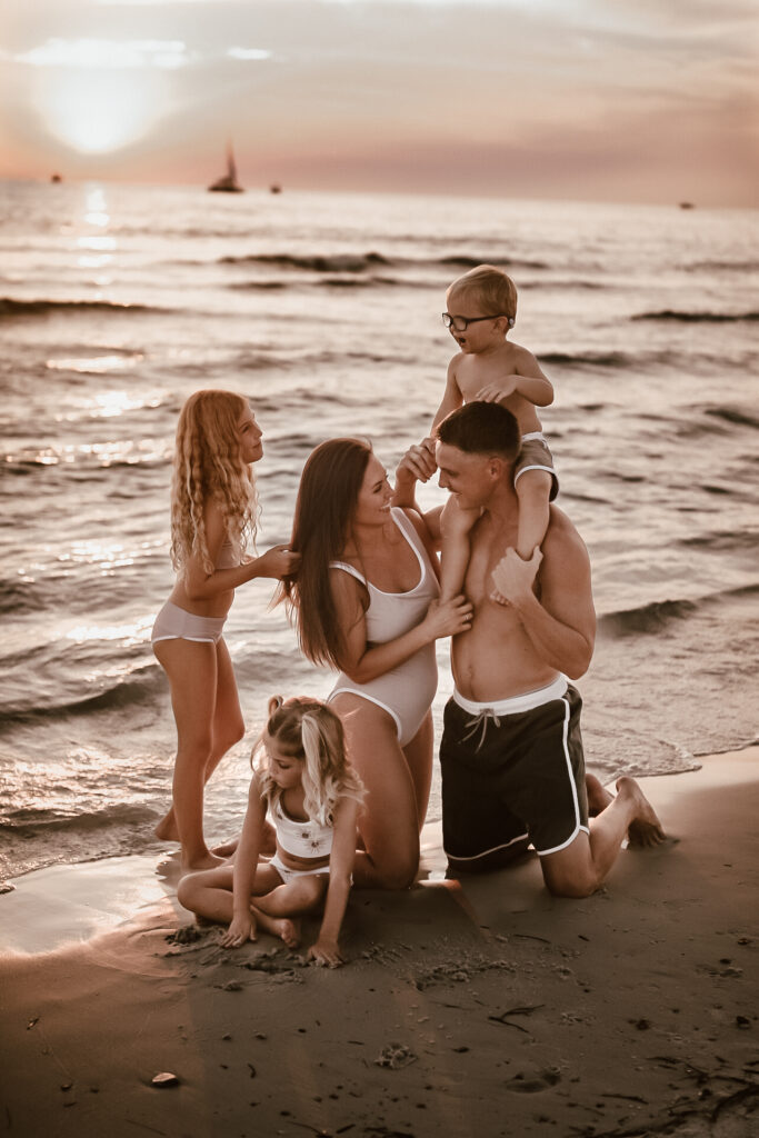 Family of 5 at St. Andrews State Park in Florida - Mom and dad kneeling in the sand looking at each other and one daughter playing in the sand at son on da' shoulders