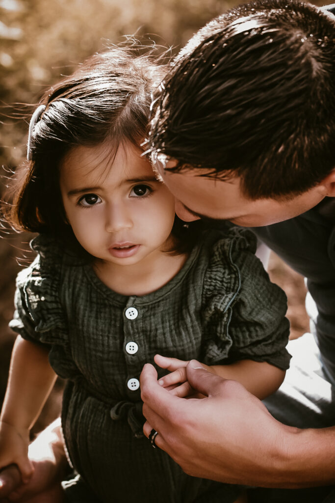 Close up of dad holding and embracing his young daughter as she looks up at the camera with a curious face