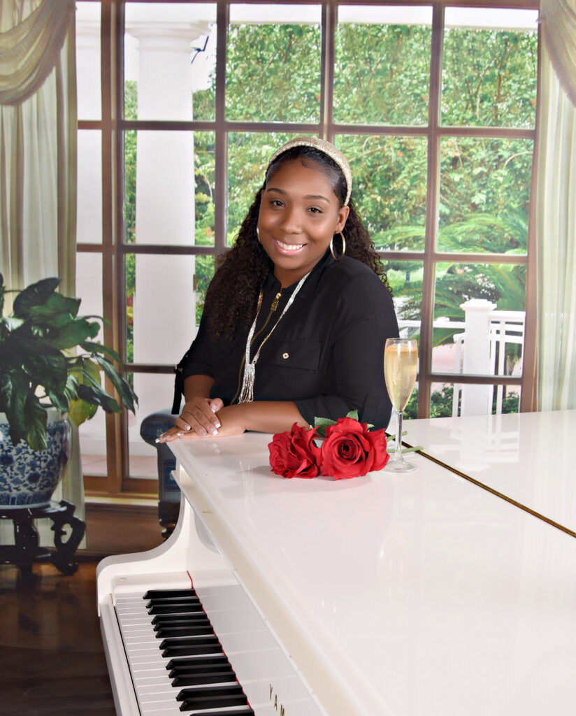 A self-portrait of Ashley, owner of Ashley Aria Photography, smiling while leaning against a white piano, with a big window in the background, displaying the beautiful scenery outside.