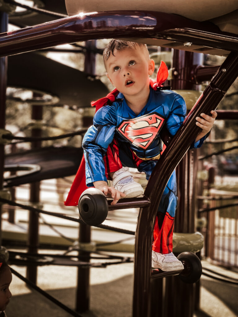 Little boy in a superman costume looking upwards while climbing at swift cantrell park