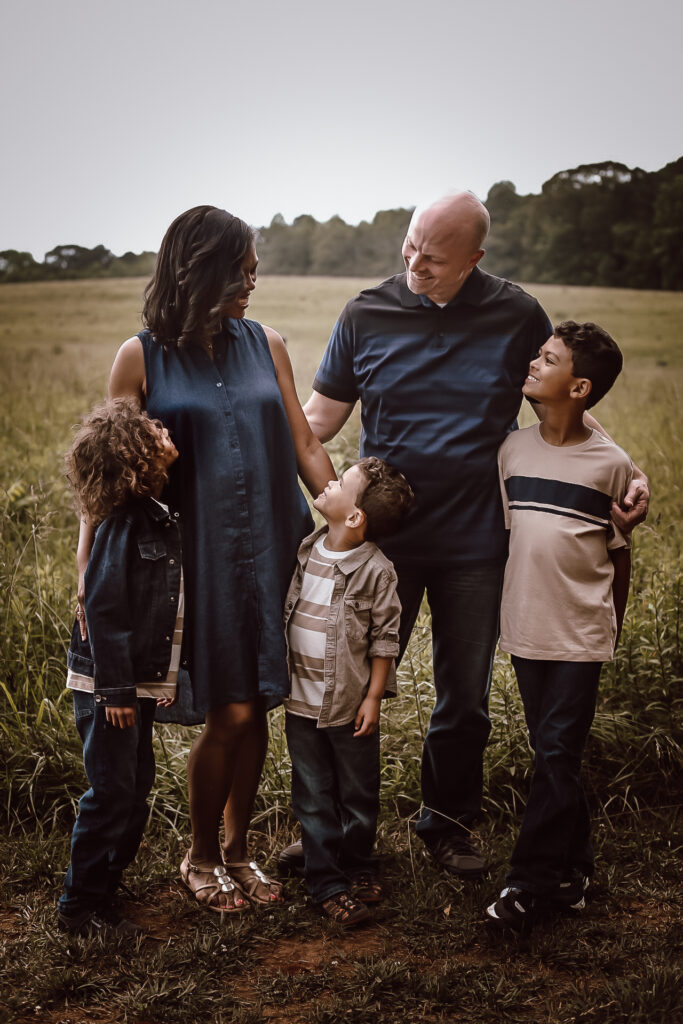 Family portrait of mom, dad, and three young boys, standing in a big open field laughing and talking with each other.