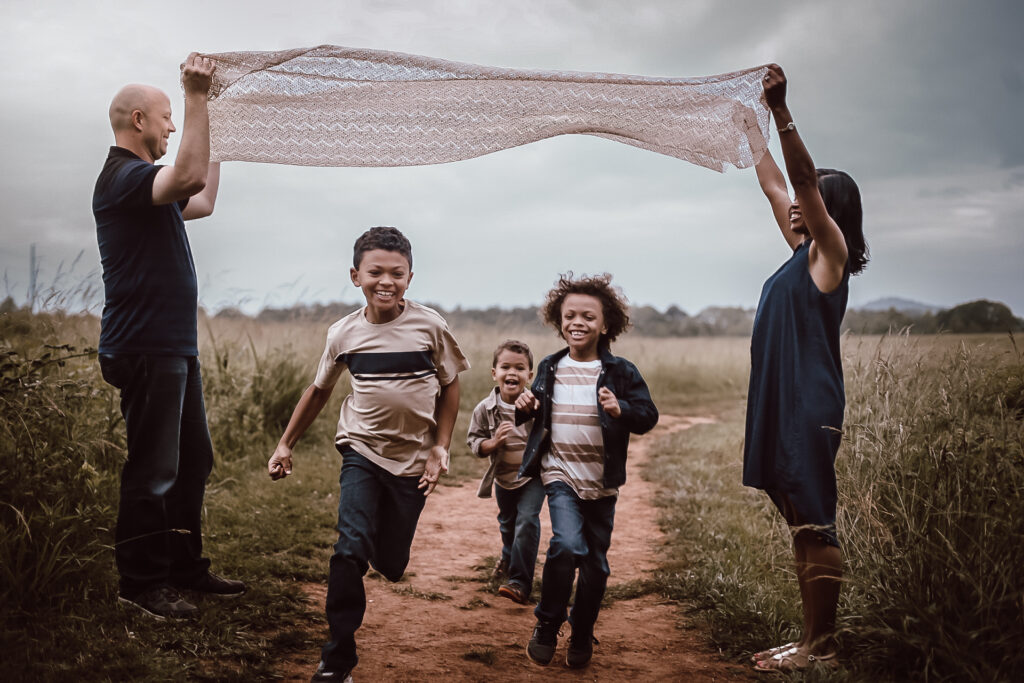 Mom and dad holding up a blanket while their three young boys run under it, laughing and playing in a big open field