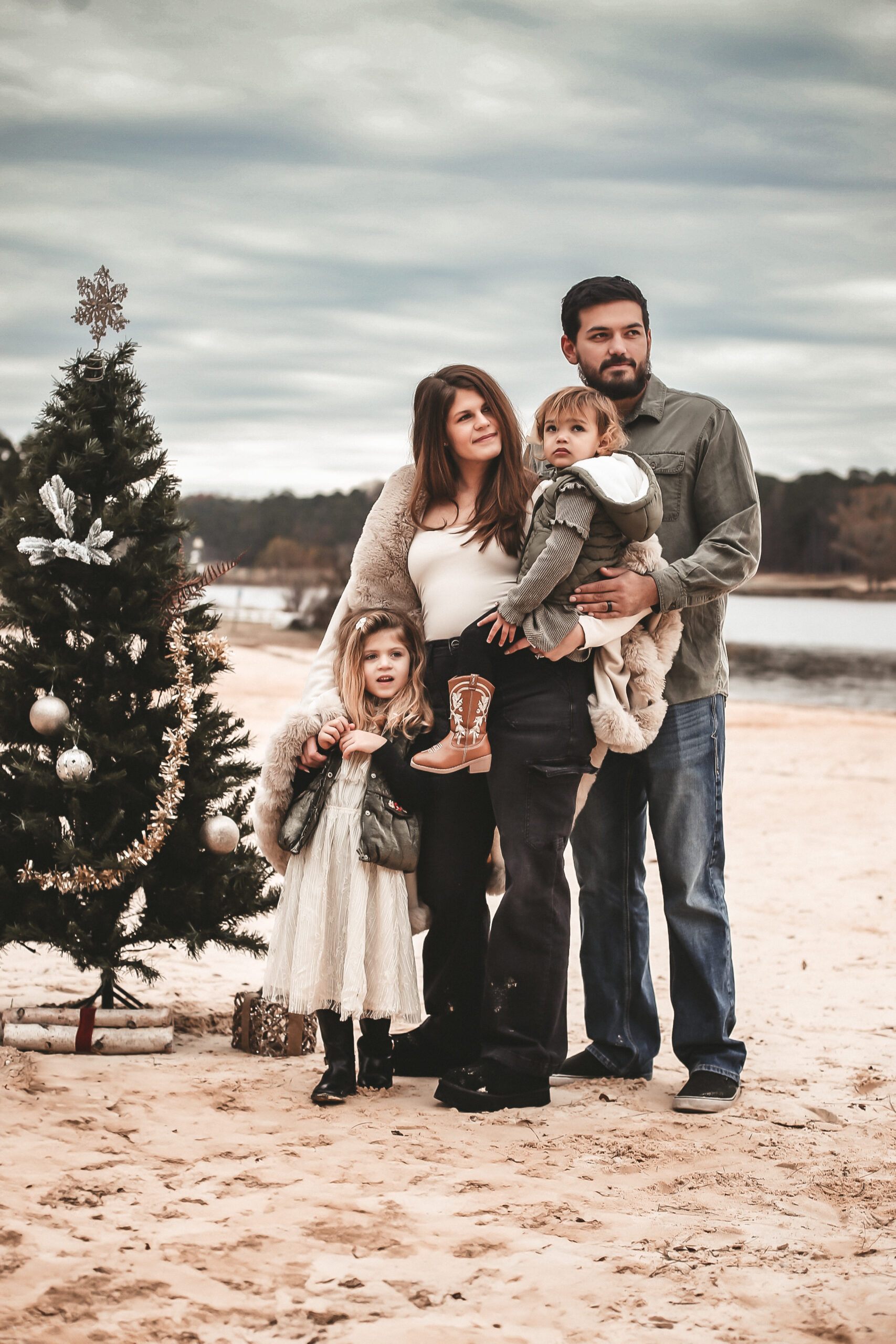 Family of 4 at the beach standing next to a Christmas tree on the sand, looking in the distance