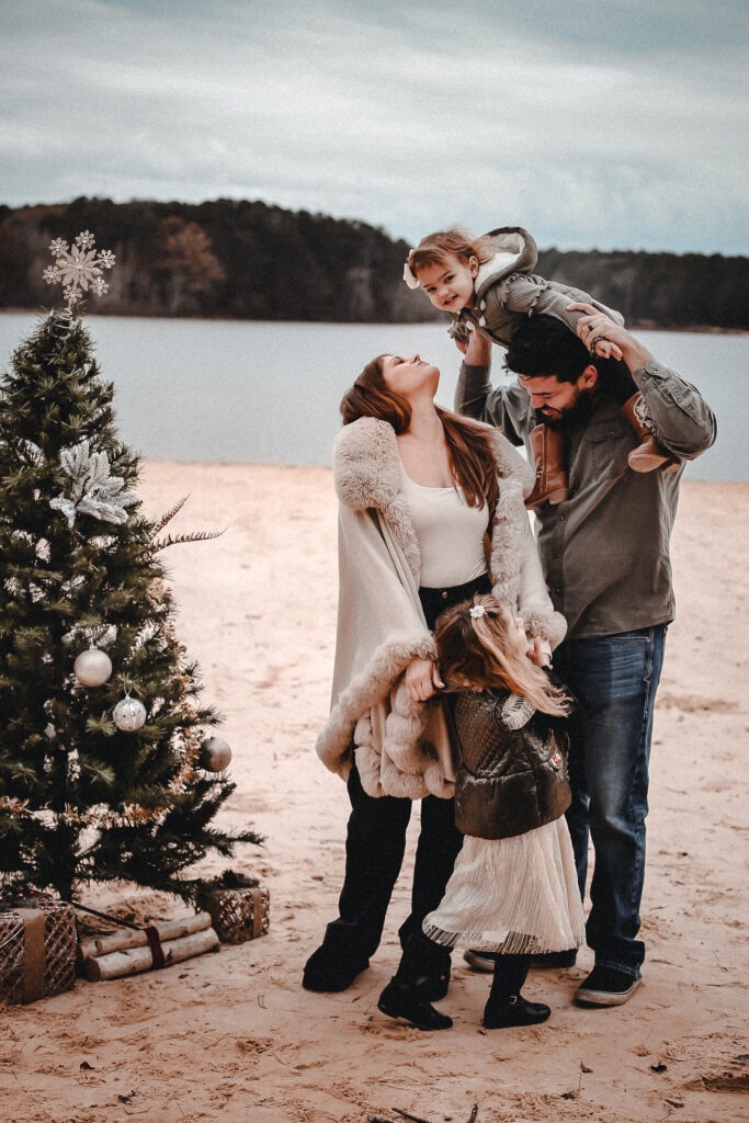 Family portrait on the beach with a Christmas tree and little girl on dads shoulders while looking at the camera and smiling and mom and sister looking up at her